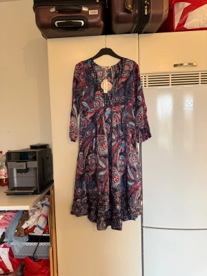 Odd Molly paisleymönstrad klänning - Snygg långklänning från Odd Molly i modellen Esemble Dress, färg French Navy. Klänningen har ett paisleymönster i blått, rött och vitt, med volangkant nedtill och trekvartsärmar. Luftig passform och v-ringad hals. Perfekt för dig som gillar boho-vibe och färgstarka prints. Storlek 2 som enligt deras storleksguide motsvarar en M. Helt ny med tags/ prislapp fortfarande kvar.  