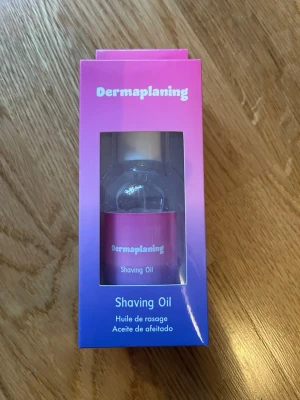 Dermaplaning Shaving Oil - Transparent rakolja för ansiktet i en liten flaska med vit kork, för att underlätta dermaplaning och ge en smidig rakning. Förpackningen är rosa och lila med instruktioner på flera språk. Oljan hjälper rakbladet att glida lätt över huden.