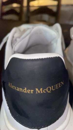 Alexander McQueen vita sneakers - Säljer ett par stilrena vita sneakers från Alexander McQueen med breda vita snören och svart häl med guldfärgad logga. Skorna har en chunky platt sula och är tillverkade i slätt skinn. Perfekta för dig som gillar lyxiga och trendiga sneakers.
