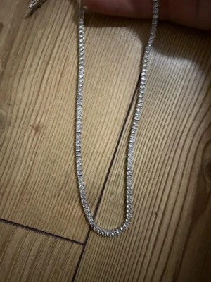 Silverfärgad moissanite tennislänk - Snygg och glittrig tennislänk från GRA med moissanite-stenar som ger maxad bling. Kedjan är äkta silver och har fyrkantiga infattningar runt varje sten för en clean och lyxig look. Perfekt för dig som vill sticka ut med något riktigt glittrigt.