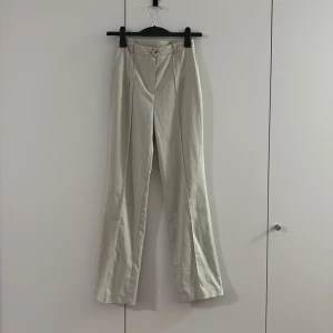 Beige kostymbyxor med raka ben - Snygga beige kostymbyxor med raka ben och pressveck framtill. Byxorna har hög midja, bälteshällor och stängs med knapp och dragkedja. Perfekta för en clean och stilren look, passar till både sneakers och boots.