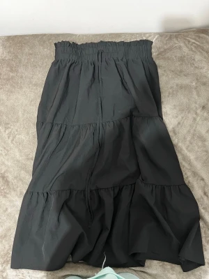 Svart långkjol med volanger - Svart långkjol med bred resår i midjan och snörning. Kjolen har flera lager med volanger som ger en skön och flowy känsla. Perfekt för dig som gillar en enkel men ändå snygg look. Materialet känns lätt och bekvämt mot huden.