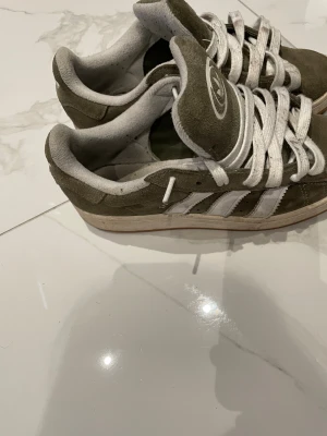 Adidas Campus sneakers i olivgrön mocka - Säljer ett par Adidas Campus sneakers i olivgrön mocka med vita detaljer och klassiska tre ränder på sidan. Skorna har rund tå, platt sula och vita snören. Adidas-logga på tungan och insidan är fodrad i ljusgrått tyg. Perfekt för dig som gillar retrostil.