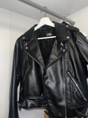 Svart oanvänd skinnjacka från Zara, S - Säljer en svart skinnjacka från Zara i storlek S. Jackan har klassisk sil med silvriga dragkedjor, bred krage och snygga detaljer som knappar och bälte vid nederkanten. Skriv om ni undrar något. Pris kan ändras