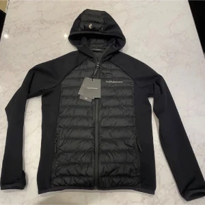  Peak Performance hybrid jacka  - Snygg svart hybrid jacka från Peak Performance med huva och hel dragkedja. Jackan har quiltad framsida och insida, två sidofickor med dragkedja och diskret logga på bröstet. Perfekt för dig som gillar stilren och sportig look. Jacka är helt ny och oanvänd med etiketter som fortfarande sitter på jackan!