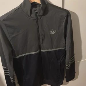Grå och svart ziptröja från Adidas - Snygg ziptröja från Adidas i grått och svart med klassiska treklövern på bröstet. Tröjan har hel dragkedja, ståkrage och diskreta ränder på ärmarna. Perfekt för chill eller träning, med praktiska fickor och sportig look.