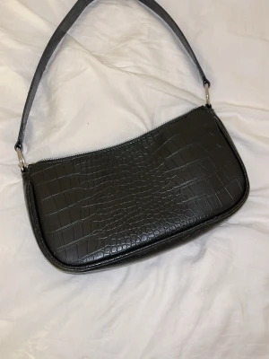 Svart axelväska i croco-imitation - Snygg svart axelväska med croco-mönstrad yta och silvrig dragkedja. Väskan har en stilren form och bred axelrem, perfekt för att lyfta vilken outfit som helst. Materialet är syntet och insidan är fodrad i svart tyg.