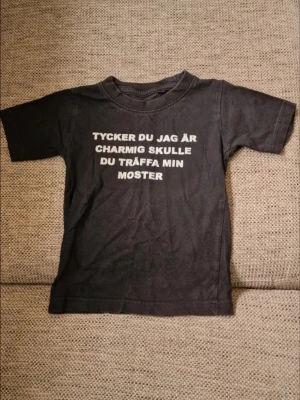 Svart t-shirt med texttryck - Svart t-shirt i mjuk bomull med vit tryckt text på framsidan: 'Tycker du jag är charmig skulle du träffa min moster'. Klassisk rund hals och korta ärmar, perfekt för dig som gillar humor och vill sticka ut lite extra. De skavanker jag har kunnat se är att det finns tvättpåverkan då färgen är lite blekt vid sömmar som halslinning. Det finns även några sprickor i trycket (se bilder). Stl 70/74