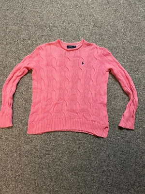 Rosa stickad tröja från Polo Ralph Lauren - Säljer äkta rosa stickad tröja från Polo Ralph Lauren i storlek XS. Tröjan har ett klassiskt kabelstickat mönster och den ikoniska broderade loggan på bröstet. Tillverkad i 100% bomull och har långa ärmar samt ribbade muddar vid ärmslut och nederkant.