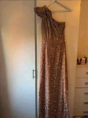 Glittrig champagnefärgad långklänning - Säljer en elegant långklänning från Miss Ord i storlek S. Klänningen är champagnefärgad med glittriga paljetter över hela tyget, har oneshoulder-design med en volangdetalj på axeln. Perfekt för dig som vill sticka ut med en glamorös look.