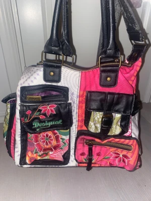 Desigual blommig axelväska med broderi - Färgstark axelväska från Desigual med blommigt broderi och flera praktiska fickor framtill. Väskan har svarta detaljer i skinnimitation, dragkedja upptill och justerbar axelrem. Mix av rosa, röd, vit och lila toner med coola mönster och broderade blommor. Axelremmen flagnar! 