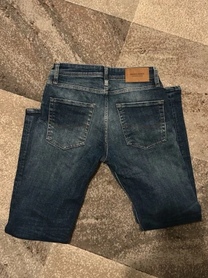Jack & Jones Regular Clark jeans blå - Säljer ett par snygga blå jeans från Jack & Jones, modell Regular Clark. Klassisk femficksdesign med slitningar och kontrastsömmar. Jeansen har rak passform och normal midja, tillverkade i slitstarkt bomullsmaterial.