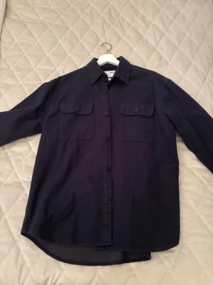 Mörkblå overshirt från Selected Homme - Säljer en stilren mörkblå overshirt från Selected Homme. Jackan har två stora bröstfickor med knappar, klassisk krage och knäppning hela vägen fram. Perfekt att slänga över en t-shirt för en chill look. Aldrig använd. Storlek S men passar M