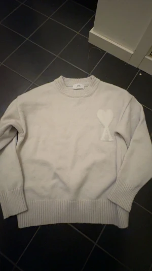  stickad tröja från Ami - Säljer en beige stickad tröja från Ami Alexandre Mattiussi med deras ikoniska hjärta-AMI Paris  på bröstet. Tröjan har rund hals, ribbade muddar och är långärmad. Perfekt för lager-på-lager under kyliga dagar.