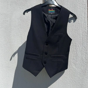 Svart kostymväst Outfitters Nation - Snygg svart kostymväst från Outfitters Nation med klassisk v-ringning och tre knappar framtill. Västen har fyra fickor fram och bröstficka och justerbart bälte bak för perfekt passform. Materialet är slätt och stilrent, perfekt till skjorta och kavaj. Storlek herr XXS och är i mycket fint skick, sparsamt använd och hel o ren. 