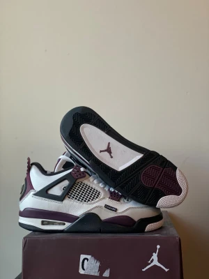 Nike Jordan 4 PSG vit och vinröd - Nike Air Jordan 4 PSG sneakers i vitt och vinrött med svarta detaljer. Skorna har meshpaneler, snörning, och Paris Saint-Germain logga på hälen. Ovandelen är i skinn och mesh, med platt sula och rund tå. Snygga detaljer som 'PANAME'-tag och vinröda accenter.