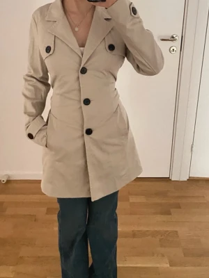 Middle length trenchcoat  - Snygg beige trenchcoat med stora svarta knappar och bälte i midjan. Jackan har klassisk krage, långa ärmar och fickor på sidorna. Perfekt för dig som gillar stilrena och tidlösa plagg. Passformen är normal och materialet känns som bomull.