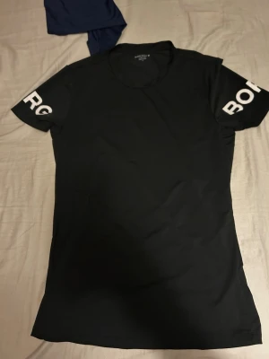 Svart t-shirt från Björn Borg - Säljer en svart t-shirt från Björn Borg med vita tryckta bokstäver på båda ärmarna. T-shirten har rund hals och korta ärmar, perfekt för en sportig och avslappnad stil. Materialet känns lätt och smidigt, vilket gör den skön att bära.