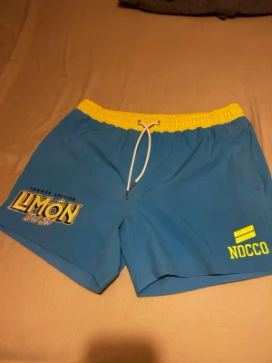 Blå NOCCO badshorts med gul midja - Säljer ett par blå badshorts från NOCCO med gul resår i midjan och vit snörning. Tryck med 'LIMÓN Dulce' och NOCCO-logga i gult på framsidan samt bakficka med logga. Perfekta för stranden eller poolen!