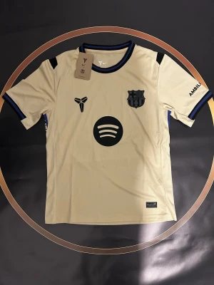 Beige FC Barcelona x Nike fotbollströja - Säljer en beige FC Barcelona x Nike fotbollströja med svarta och blå detaljer. Tröjan har tryck med Spotify-logga, Kobe Bryant-symbol, klubbmärke och nummer 10 med Lamine Yamal på ryggen. Materialet är lätt polyester och har ribbad krage och ärmslut.