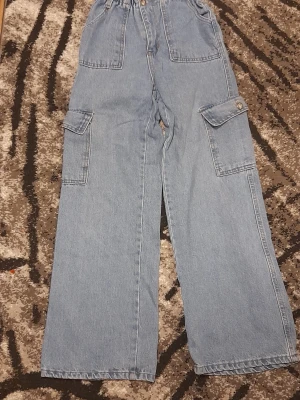 Blå cargopants i jeansmaterial - Säljer ett par ljusblå cargopants i jeansmaterial med breda ben och stora fickor på sidorna. Byxorna har normal passform och klassisk midja, samt knappstängning på fickorna. Perfekt för dig som gillar streetwear och praktiska detaljer. Jättefina baggy cargopants men ör för små