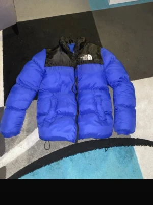 Blå pufferjacka från The North Face - Blå och svart pufferjacka från The North Face med broderad logga på bröstet. Jackan har dragkedja framtill, hög krage och två stora fickor. Perfekt för kalla dagar och har en riktigt schysst streetstil.