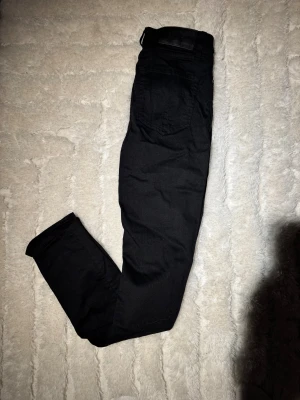 Svarta jeansbyxor slim fit från tiger of Sweden 29/32 - Svarta jeansbyxor med slim fit-passning och klassisk femficksdesign. Byxorna har en stilren look med svarta sömmar och en diskret svart läderpatch vid linningen. Perfekt för dig som gillar en clean och enkel stil. Skriv om du vill ha mer mått