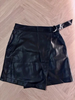 Svart omlottkjol Twist & Tango - Snygg svart kort omlottkjol från Twist & Tango i skinnimitation med coolt bälte och metallspänne i midjan. 