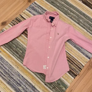 Rosa skjorta från Ralph Lauren M - Pris kan diskuteras. Klassisk rosa skjorta från Ralph Lauren med lång ärm, knappar framtill och broderad logga på bröstet. Skjortan är tillverkad i mjuk bomull med lite stretch för extra komfort. Perfekt för en clean och stilren look.OBS barn storlek