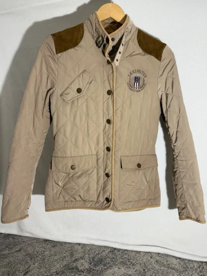 Beige quiltad jacka från Lexington - Snygg beige quiltad jacka från Lexington med bruna mockadetaljer på axlarna. Jackan har ståkrage med knappar, tre fickor framtill och Lexington-logga på bröstet. Materialet är polyester och nylon, med foder i polyester. Perfekt för dig som gillar klassisk stil.