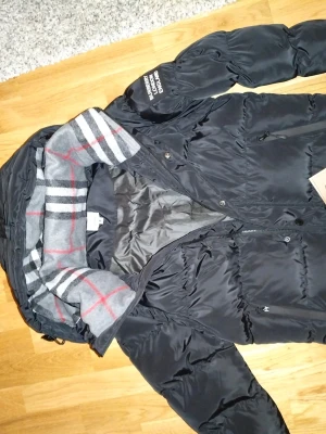 Svart pufferjacka/väst från Burberry - Svart pufferjacka från Burberry med klassiskt rutigt foder i grått, vitt och rött. Jackan har dragkedja och tryckknappar framtill, samt flera praktiska fickor. Materialet är polyester och dun, perfekt för kalla dagar. Man kan ta bort ärmarna och göra jackan till en väst! Snyggt Burberry-tryck på ärmen.