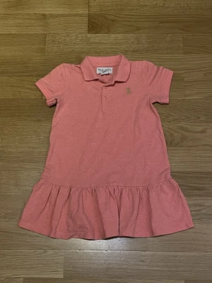 Rosa klänning från Holly & White för barn - Säljer en söt rosa klänning från Holly & White med krage och korta ärmar för barn!  Klänningen har en volang nedtill och knappslå vid halsen. Perfekt för dig som gillar en enkel och fräsch stil.