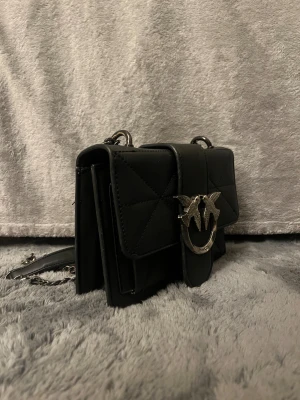 Svart quiltad axelväska från Pinko - Säljer en svart quiltad axelväska från Pinko med snyggt silverfärgat spänne med fåglar framtill. Väskan har kedjerem med läderdetalj och praktiska fack. Perfekt storlek för mobilen och dina viktigaste grejer.