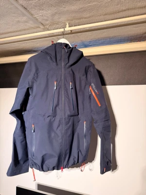 Stellar equipment shell jacket  - Mörkblå skaljacka från Stellar Equipment med alla funktioner man kan behöva för skidåkning eller andra friluftsaktiviteter.  Jag har huvudsakligen använt den för utförsåkning men funkar utmärkt till allt möjligt. Den är i utmärkt skick förutom ett brännmärke på utsidan som jag fick av ett fall på en rail (se bild) märks inte så mycket när man har på sig den. Då den bara är ytlig och inte gått igenom hela skalet går fukten inte igenom, visar bara att åkaren är seriös i backen😅  Jackan är fullt ut