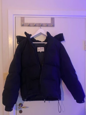 Svart pufferjacka från Levi's - Svart pufferjacka från Levi's med huva och dragkedja framtill. Jackan har en loose passform och snörning vid nederkanten. Perfekt för kalla dagar och har en stilren look som funkar till många outfits.