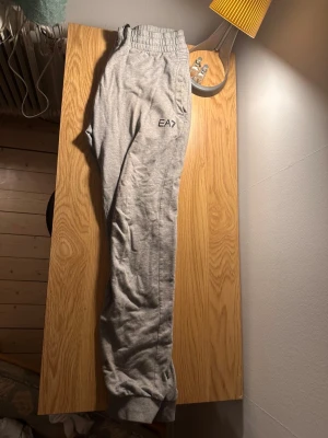 Grå mjukisbyxor från EA7 Emporio Armani - Säljer ett par grå mjukisbyxor från EA7 Emporio Armani med elastisk midja och snörning. Byxorna har fickor på sidorna och EA7-logga tryckt på ena benet. Tillverkade i mjukt bomullsmaterial för maximal komfort.
