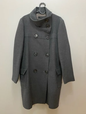Grå ullkappa från Zara Woman - Snygg grå kappa från Zara Woman i ullmix med dubbelknäppning och hög krage. Klassisk rak modell med stora fickor och stilrena knappar framtill. Perfekt för dig som vill ha en clean och tidlös look till höst och vinter.