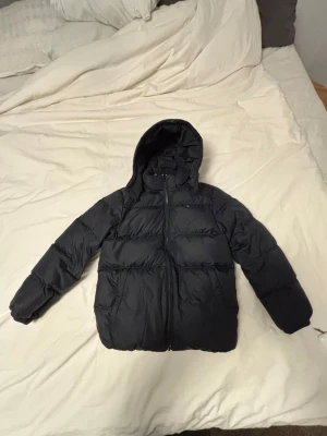 Svart pufferjacka från Tommy Hilfiger - Svart pufferjacka från Tommy Hilfiger med huva och broderad logga på insidan. Jackan har dragkedja framtill, två sidofickor och klassisk quiltad design. Materialet är syntet och jackan är perfekt för kalla dagar. Snygg och stilren look med diskret logga på bröstet. Endast använd en vintersäsong ! Köpt för 2000kr!