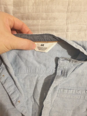 Ljusblå skjorta från H&M med knappar - Snygg ljusblå skjorta från H&M med diskret mönster och klassisk krage. Skjortan har långa ärmar, bröstficka och knappar framtill i kontrastfärg. Perfekt för dig som gillar enkel och stilren look.