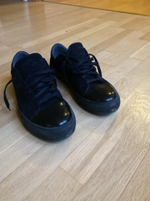Svarta sneakers från Sneaky Steve - Snygga svarta sneakers från Sneaky Steve med mocka på sidorna och blankt skinn på tå och häl. Skorna har rund tå, platt sula och klassisk snörning. Perfekta för dig som gillar stilrena och enkla skor med en cool touch.
