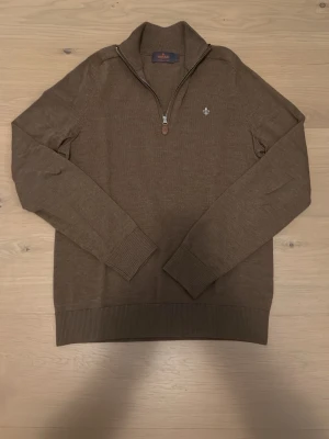 Brun merinoulltröja från Morris - Säljer en brun långärmad stickad tröja från Morris i 100% merinoull. Tröjan har half zip, ribbade muddar och en liten broderad logga på bröstet. Perfekt för dig som gillar klassisk stil och vill ha något mjukt och bekvämt.
