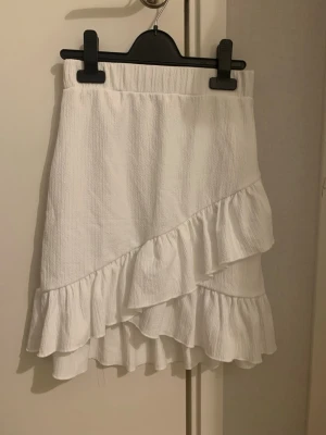 Vit volangkjol från nelly - Supersöt vit kjol från Nelly i storlek XS. Kjolen har volanger nere vid kanten och ett elastiskt midjeband för skön passform. Är oanvänd i nytt skick. Materialet är lätt och luftigt, perfekt för varma sommardagar och för dig som vill ha en fin fräsch look.