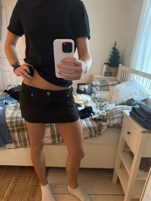 Svart kortkjol med fickor och knappar - Säljer en svart kortkjol med flera fickor och snygga silverfärgade knappar. Kjolen har bälteshällor och coola detaljer som sömmar och nitar. Materialet känns som bomull och har en lite robust känsla. Perfekt för dig som gillar streetstyle och utility-vibe.