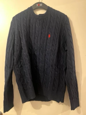 Marinblå stickad tröja Polo Ralph Lauren - Säljer en marinblå kabelstickad tröja från Polo Ralph Lauren i storlek M. Tröjan har ett klassiskt rött broderat logotyp på bröstet, rund hals och långa ärmar. Materialet är mjukt och stickat, perfekt för kyliga dagar.