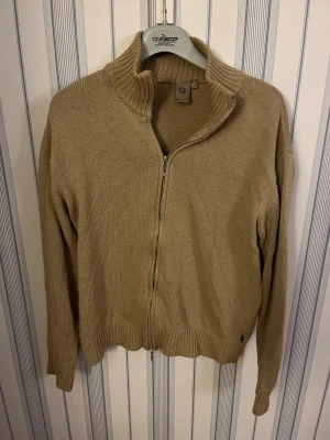 Beige stickad tröja full zip - Säljer en beige stickad kofta med hög krage och hel dragkedja framtill. Koftan har ribbade muddar vid ärmslut och nederkant, samt långärmad passform. Perfekt för lager-på-lager under kyliga dagar. Tröjan är large men sitter som small.