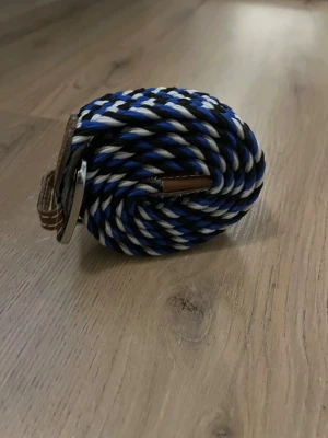Flättat bälte - Säljer ett snyggt bälte med randigt mönster i blått, svart och vitt. Bältet har en modern look med twistad design och detaljer i brunt skinn vid spännet. Perfekt för dig som vill sticka ut med färg och stil.