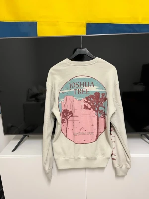 Beige sweatshirt Joshua Tree - Säljer en beige sweatshirt med tryck från Joshua Tree National Park. Tröjan har rund hals, långa ärmar med tryck i rosa och blått samt text längs ena ärmen. Mjuk bomullskänsla och relaxed fit, perfekt för chill dagar.