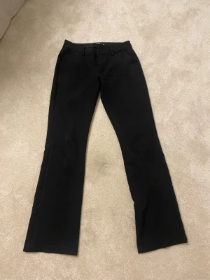 Svarta bootcut kostymbyxor - Snygga svarta kostymbyxor med bootcut-snitt och klassiska hällor för bälte. Byxorna har en stilren look och är perfekta för dig som gillar en tidlös och clean stil. Materialet känns mjukt och har en lätt stretch för extra komfort.