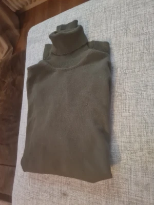 Mörkgrön polotröja från JL - Snygg mörkgrön polotröja från JL med klassisk hög krage. Tröjan är stickad och har en enkel, stilren design utan mönster eller detaljer. Perfekt för dig som gillar en clean look och vill hålla dig varm under kyliga dagar.