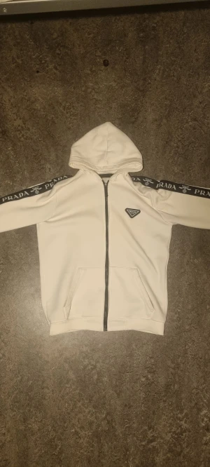 Vit Prada Hoodie - Vit hoodie från Prada med svarta band längs ärmarna där det står 'PRADA MILANO'. Passar mig lite stor som är (175cm 70kg) men den skulle även passa ngn lite större.Tröjan har huva, dragkedja framtill och två fickor. På bröstet sitter en svartvit Prada-logga. Materialet känns mjukt och är troligen bomull.
